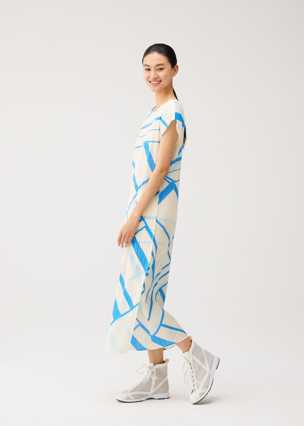 A woman smiles while wearing the FOLD LINE white round-neck midi dress with blue geometric patterns and French sleeves from PLEATS PLEASE ISSEY MIYAKE, paired with white high-top sneakers, standing against a plain light background. view 8 of 8