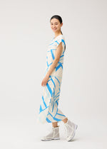 A woman smiles while wearing the FOLD LINE white round-neck midi dress with blue geometric patterns and French sleeves from PLEATS PLEASE ISSEY MIYAKE, paired with white high-top sneakers, standing against a plain light background.