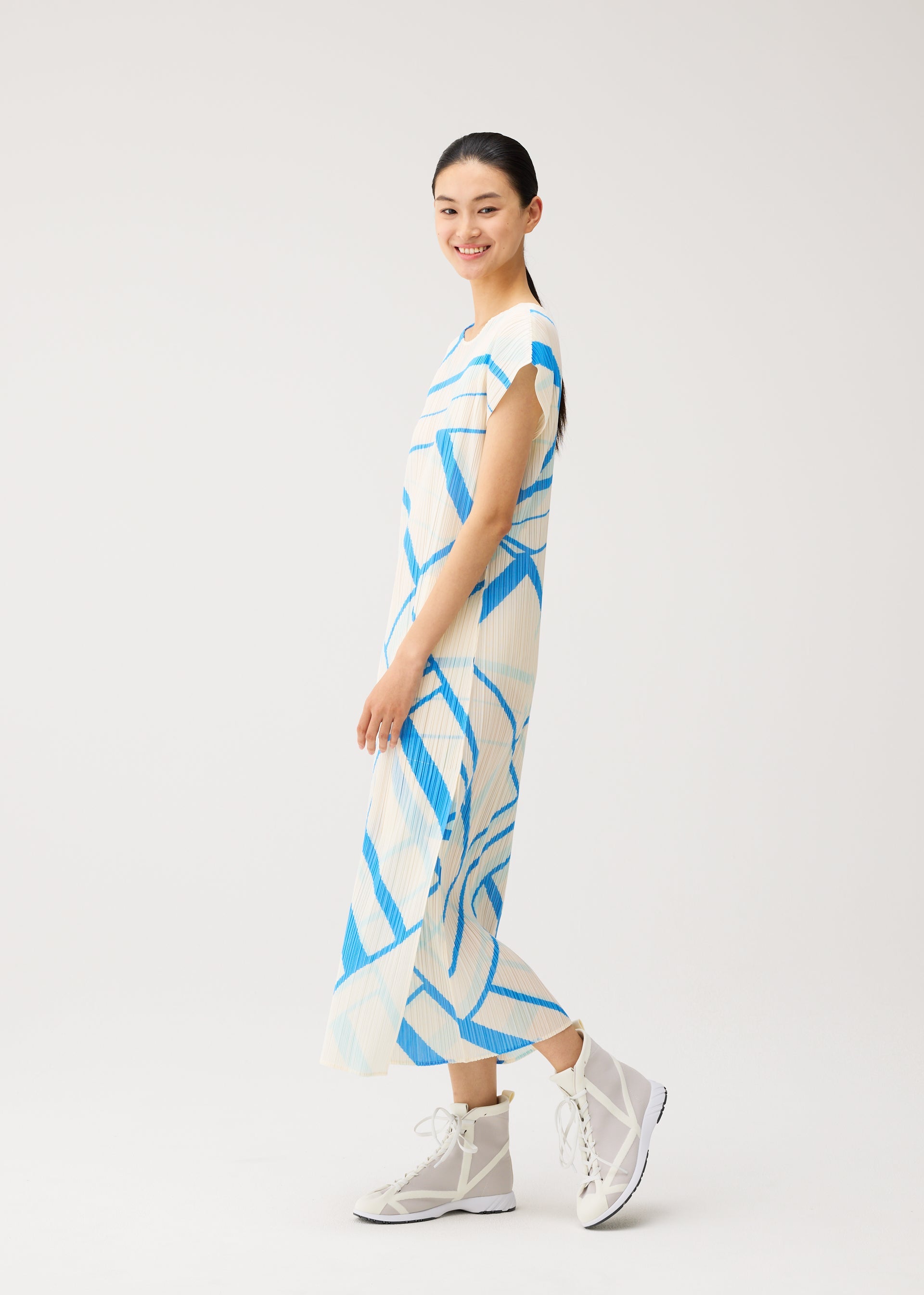 Zoomed view A woman smiles while wearing the FOLD LINE white round-neck midi dress with blue geometric patterns and French sleeves from PLEATS PLEASE ISSEY MIYAKE, paired with white high-top sneakers, standing against a plain light background.