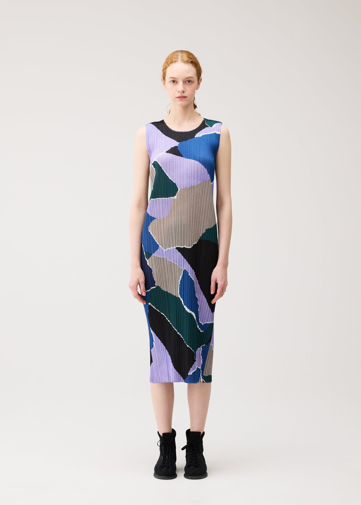 A woman stands against a plain background, wearing the “SURPRISE!” sleeveless, round-neck dress by PLEATS PLEASE ISSEY MIYAKE, featuring an abstract print in purple, green, black, and tan, paired with black lace-up shoes. view 4 of 7