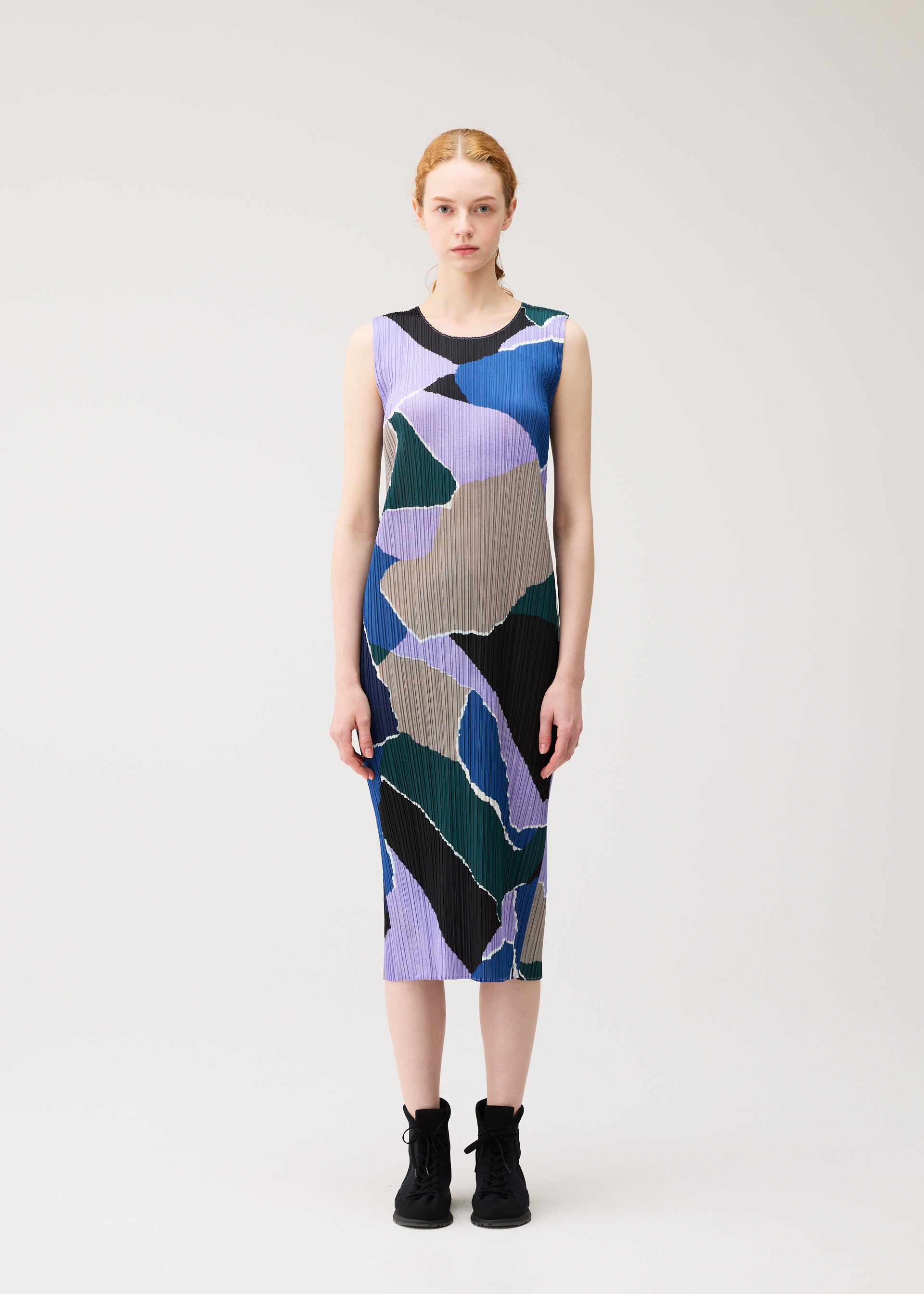 Zoomed view A woman stands against a plain background, wearing the “SURPRISE!” sleeveless, round-neck dress by PLEATS PLEASE ISSEY MIYAKE, featuring an abstract print in purple, green, black, and tan, paired with black lace-up shoes.