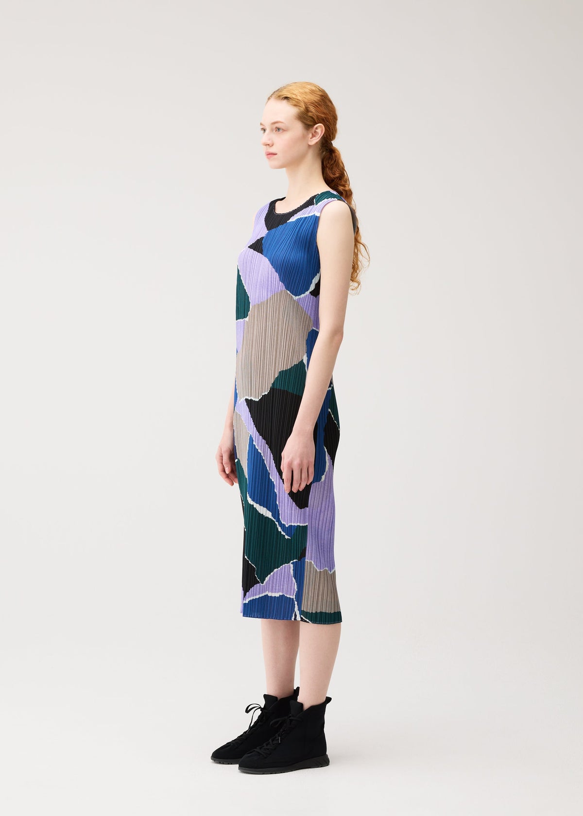 A woman with long, curly red hair stands in profile against a light background, wearing the SURPRISE! dress by PLEATS PLEASE ISSEY MIYAKE—a sleeveless, knee-length piece featuring abstract blue, green, beige, and purple patterns—and black ankle boots. view 5 of 7