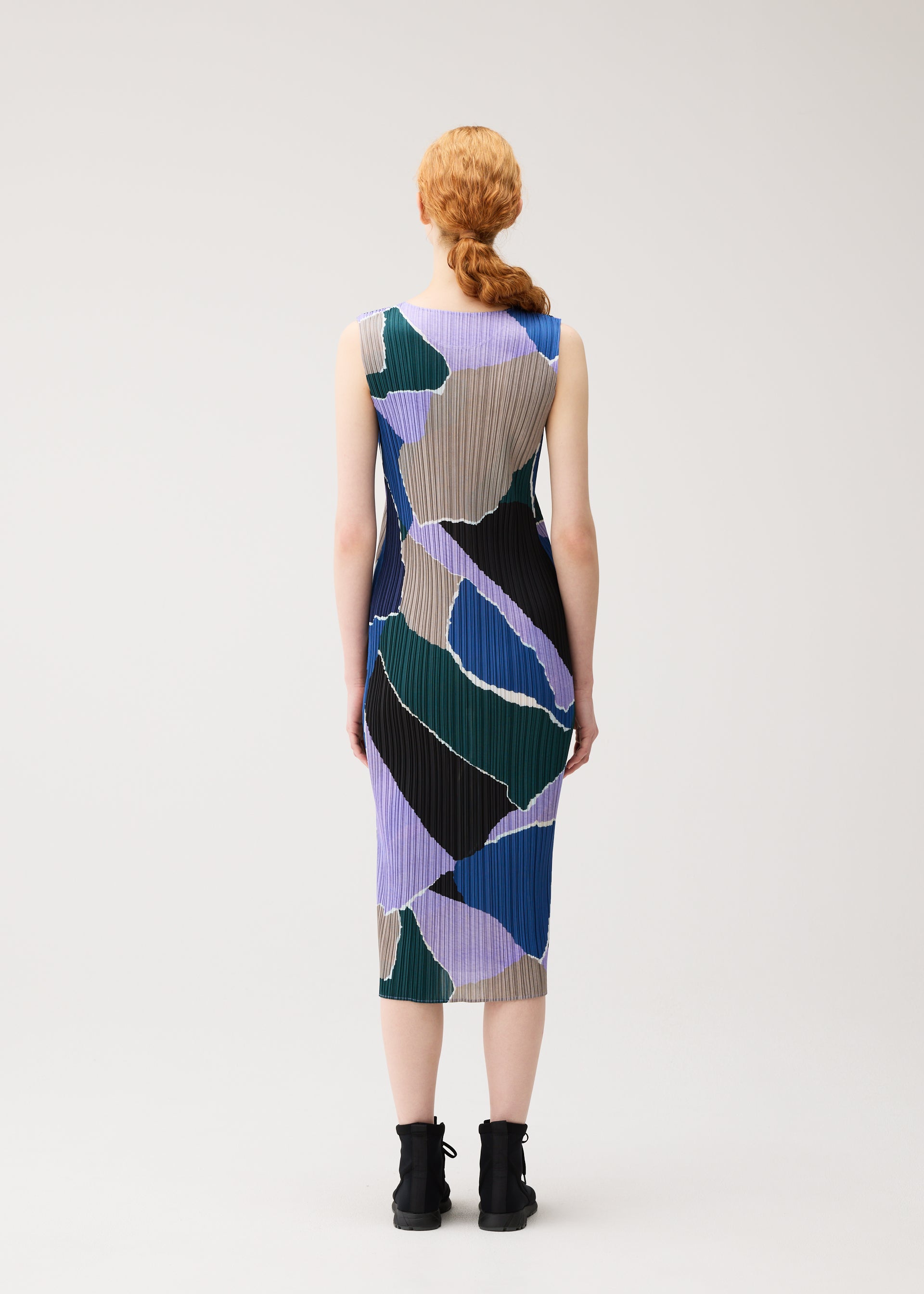 Zoomed view A person with long red hair in a low braid is seen from behind, wearing the SURPRISE! dress by PLEATS PLEASE ISSEY MIYAKE—a sleeveless, knee-length piece featuring an abstract pattern in blue, green, tan, and black—paired with black ankle boots, standing against a plain white background.