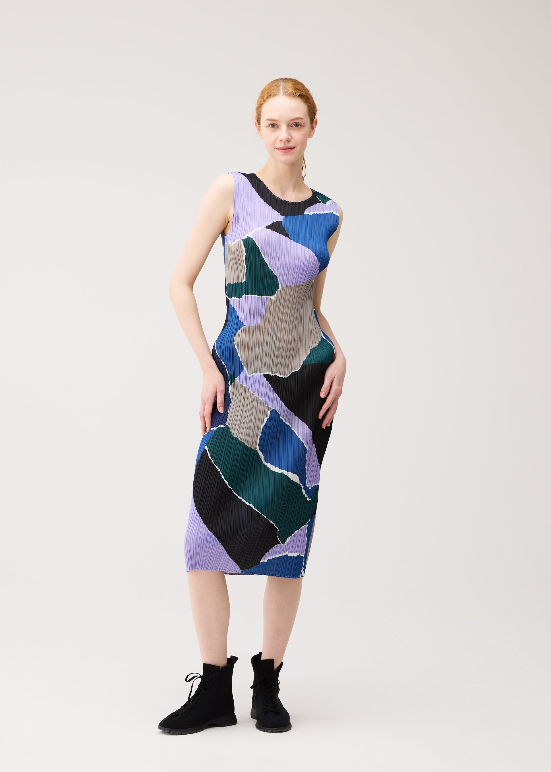 Zoomed view A woman poses in the SURPRISE! dress by PLEATS PLEASE ISSEY MIYAKE—a sleeveless, round-neck style featuring a vibrant abstract pattern in shades of purple, blue, green, black, and gray—paired with black ankle boots against a plain background.