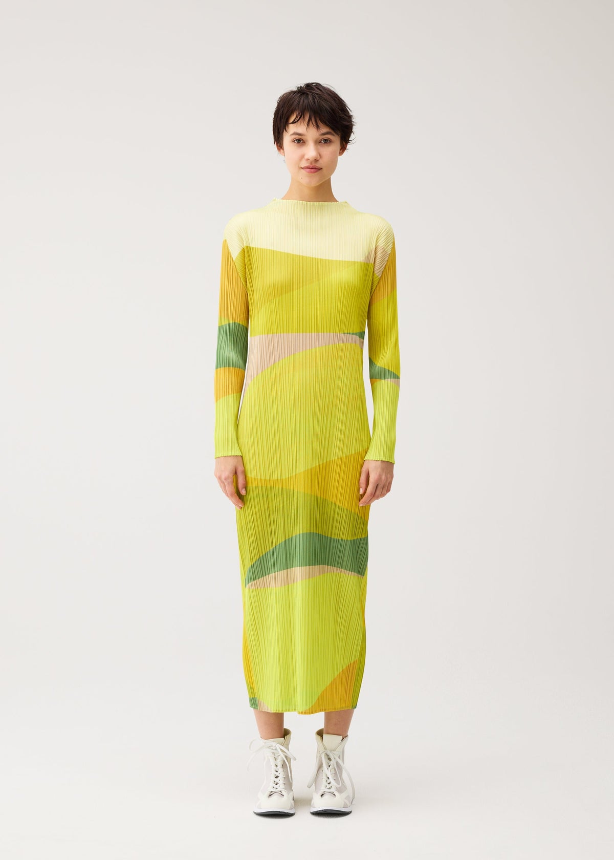 A person stands facing forward in the ORBIT dress by PLEATS PLEASE ISSEY MIYAKE—long-sleeved, yellow and green geometric print—paired with white lace-up boots against a plain light background. view 4 of 7