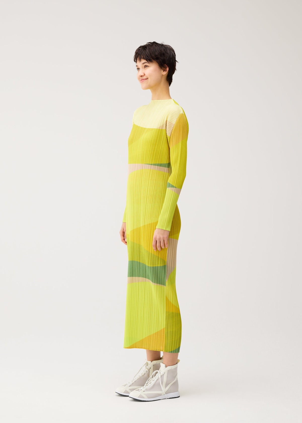 A person stands in profile wearing the ORBIT long-sleeved, ankle-length yellow dress with geometric patterns by PLEATS PLEASE ISSEY MIYAKE and white high-top sneakers against a plain light background. view 5 of 7