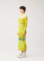 A person stands in profile wearing the ORBIT long-sleeved, ankle-length yellow dress with geometric patterns by PLEATS PLEASE ISSEY MIYAKE and white high-top sneakers against a plain light background.