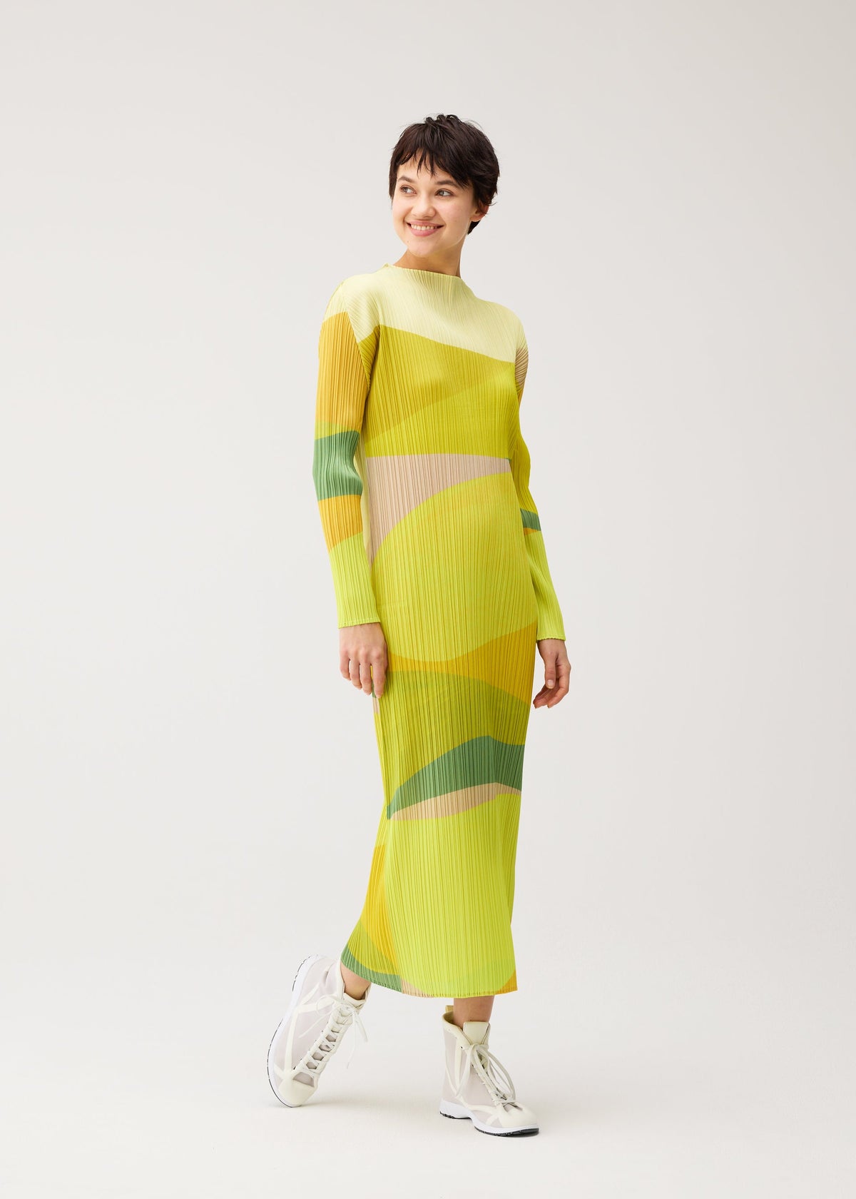 Zoomed view A smiling person with short dark hair wears the ORBIT dress by PLEATS PLEASE ISSEY MIYAKE—long, high-necked, yellow with green and orange abstract patterns—paired with white high-top sneakers on a light background.
