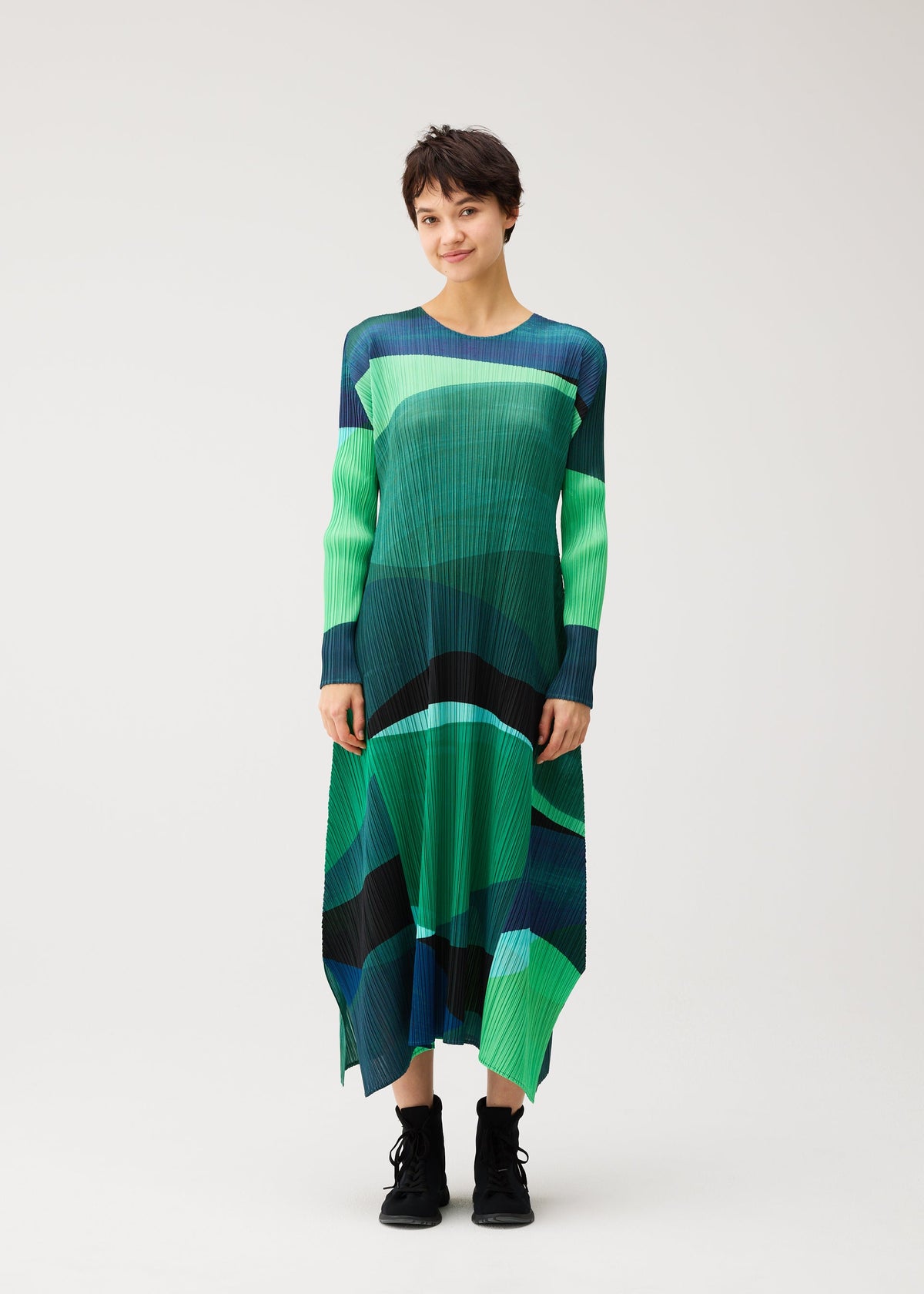 A person in the ORBIT dress by PLEATS PLEASE ISSEY MIYAKE—green and blue abstract pleated, round-neck, long-sleeved—smiles against a plain background, wearing black ankle boots. view 5 of 8