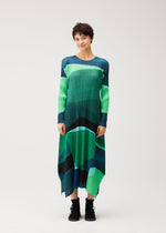 A person in the ORBIT dress by PLEATS PLEASE ISSEY MIYAKE—green and blue abstract pleated, round-neck, long-sleeved—smiles against a plain background, wearing black ankle boots.