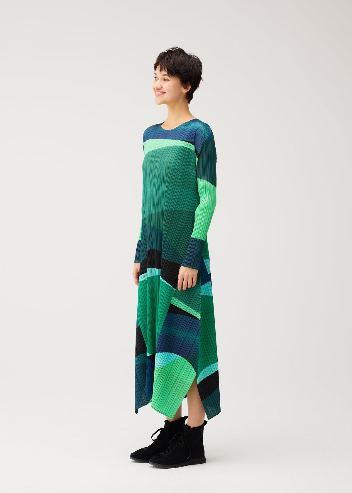 A person stands smiling against a plain white background, wearing the ORBIT dress from PLEATS PLEASE ISSEY MIYAKE, featuring a green and blue geometric pattern and paired with black ankle boots. view 6 of 8