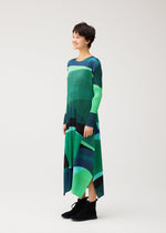 A person stands smiling against a plain white background, wearing the ORBIT dress from PLEATS PLEASE ISSEY MIYAKE, featuring a green and blue geometric pattern and paired with black ankle boots.