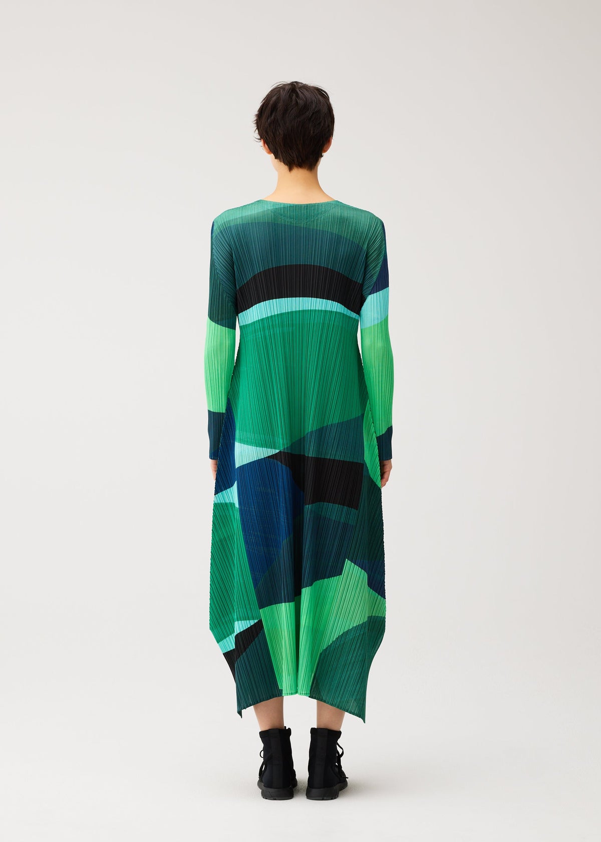 Zoomed view A person with short dark hair, seen from behind, wears the ORBIT dress by PLEATS PLEASE ISSEY MIYAKE in green and blue color-blocks with black ankle boots against a plain light background.