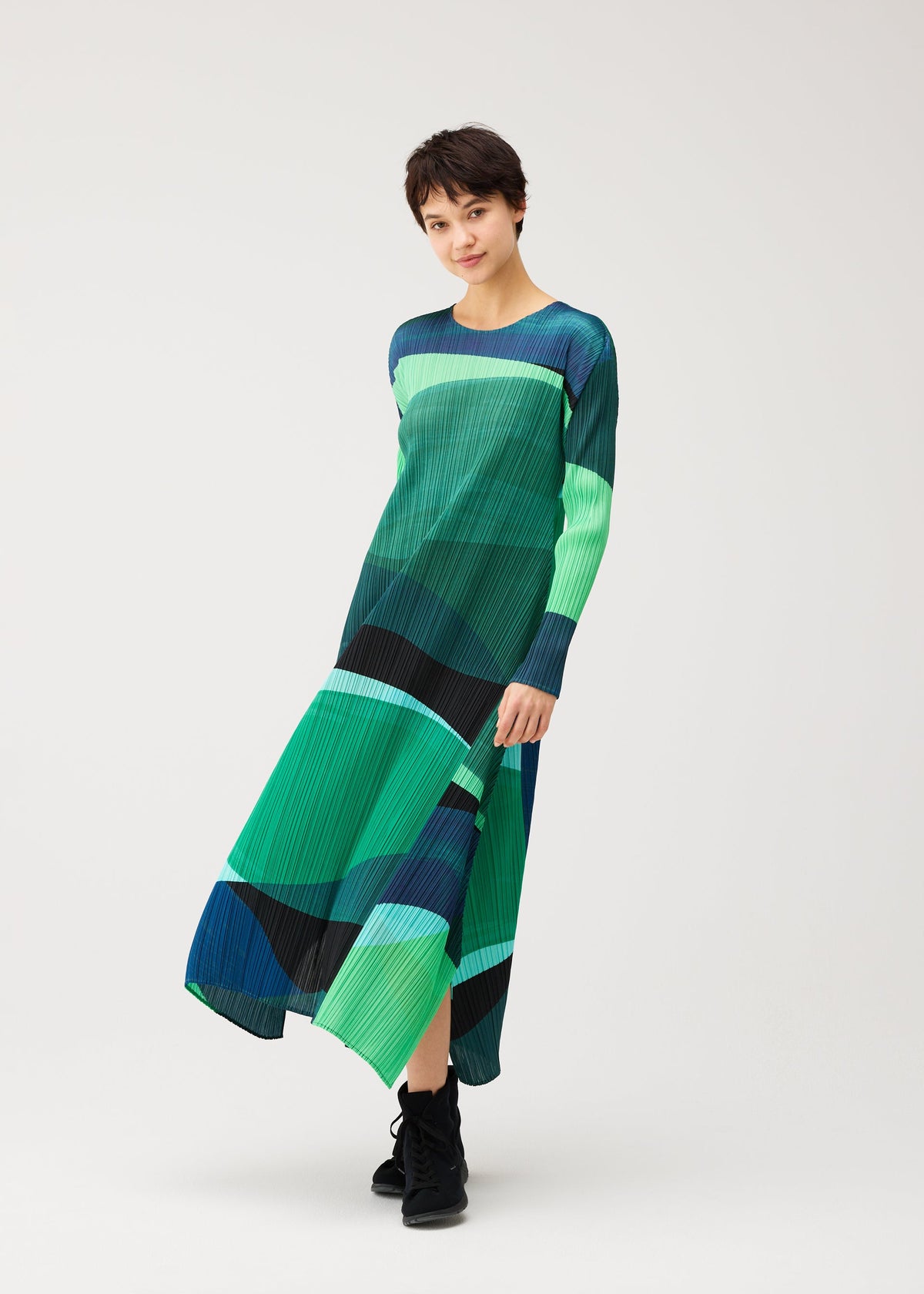 A woman wears the ORBIT dress by PLEATS PLEASE ISSEY MIYAKE, featuring green, blue, and black geometric patterns, standing against a white background and smiling slightly at the camera. view 8 of 8