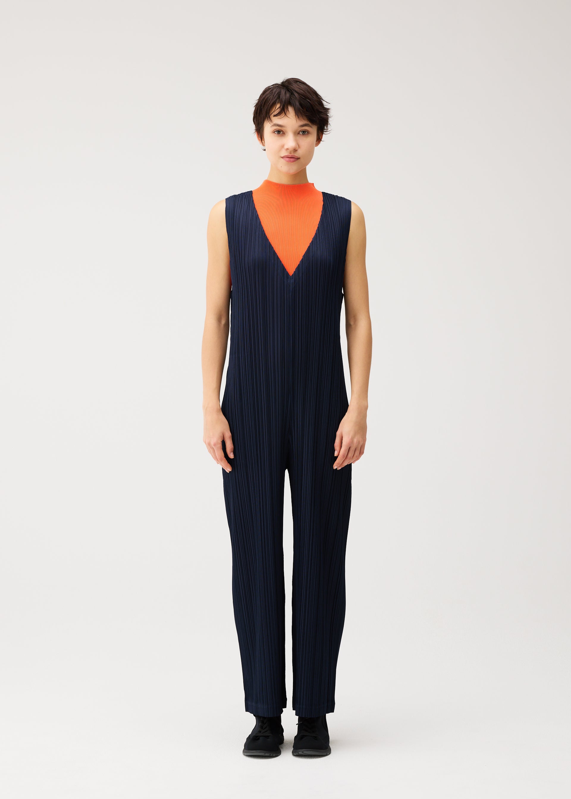 Zoomed view A person stands against a plain light background wearing PLEATS PLEASE ISSEY MIYAKE’s THICKER BOTTOMS 2—a sleeveless navy V-neck jumpsuit with pockets and thick fabric—styled over an orange high-neck top and black shoes.