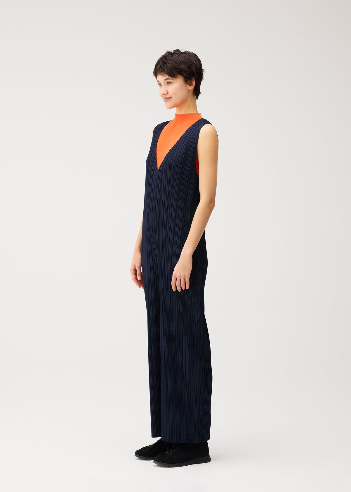A person stands in PLEATS PLEASE ISSEY MIYAKE’s THICKER BOTTOMS 2—a navy blue V-neck jumpsuit crafted from thick fabric—layered over an orange top, with short dark hair, black shoes, and a neutral background. view 5 of 7