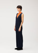A person stands in PLEATS PLEASE ISSEY MIYAKE’s THICKER BOTTOMS 2—a navy blue V-neck jumpsuit crafted from thick fabric—layered over an orange top, with short dark hair, black shoes, and a neutral background.