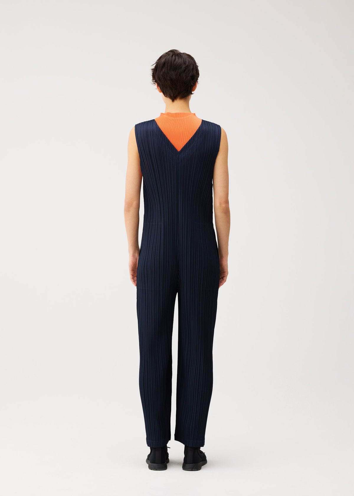 A person stands facing away, wearing the PLEATS PLEASE ISSEY MIYAKE THICKER BOTTOMS 2 jumpsuit—a sleeveless, navy blue, pleated V-neck design crafted from thick fabric—featuring a deep V-shaped back that reveals an orange underlayer. The background is plain and light-colored. view 6 of 7