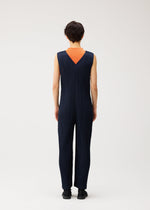 A person stands facing away, wearing the PLEATS PLEASE ISSEY MIYAKE THICKER BOTTOMS 2 jumpsuit—a sleeveless, navy blue, pleated V-neck design crafted from thick fabric—featuring a deep V-shaped back that reveals an orange underlayer. The background is plain and light-colored.