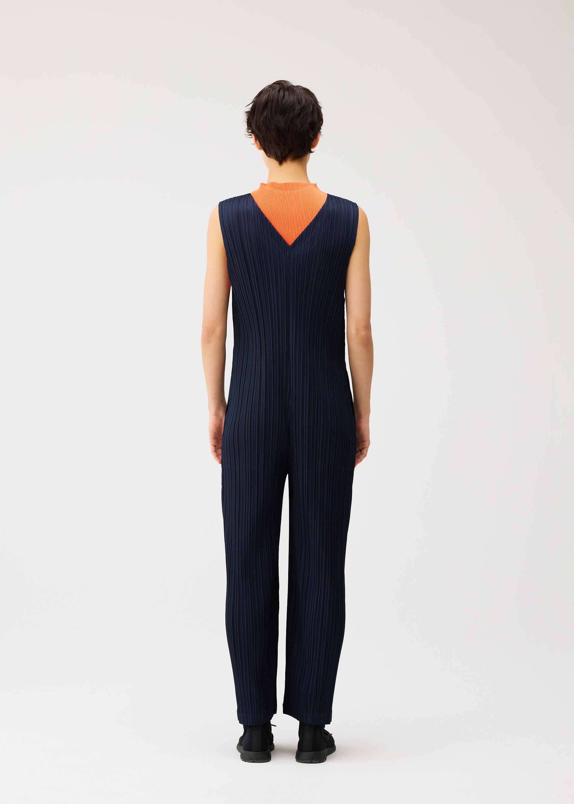 Zoomed view A person stands facing away, wearing the PLEATS PLEASE ISSEY MIYAKE THICKER BOTTOMS 2 jumpsuit—a sleeveless, navy blue, pleated V-neck design crafted from thick fabric—featuring a deep V-shaped back that reveals an orange underlayer. The background is plain and light-colored.