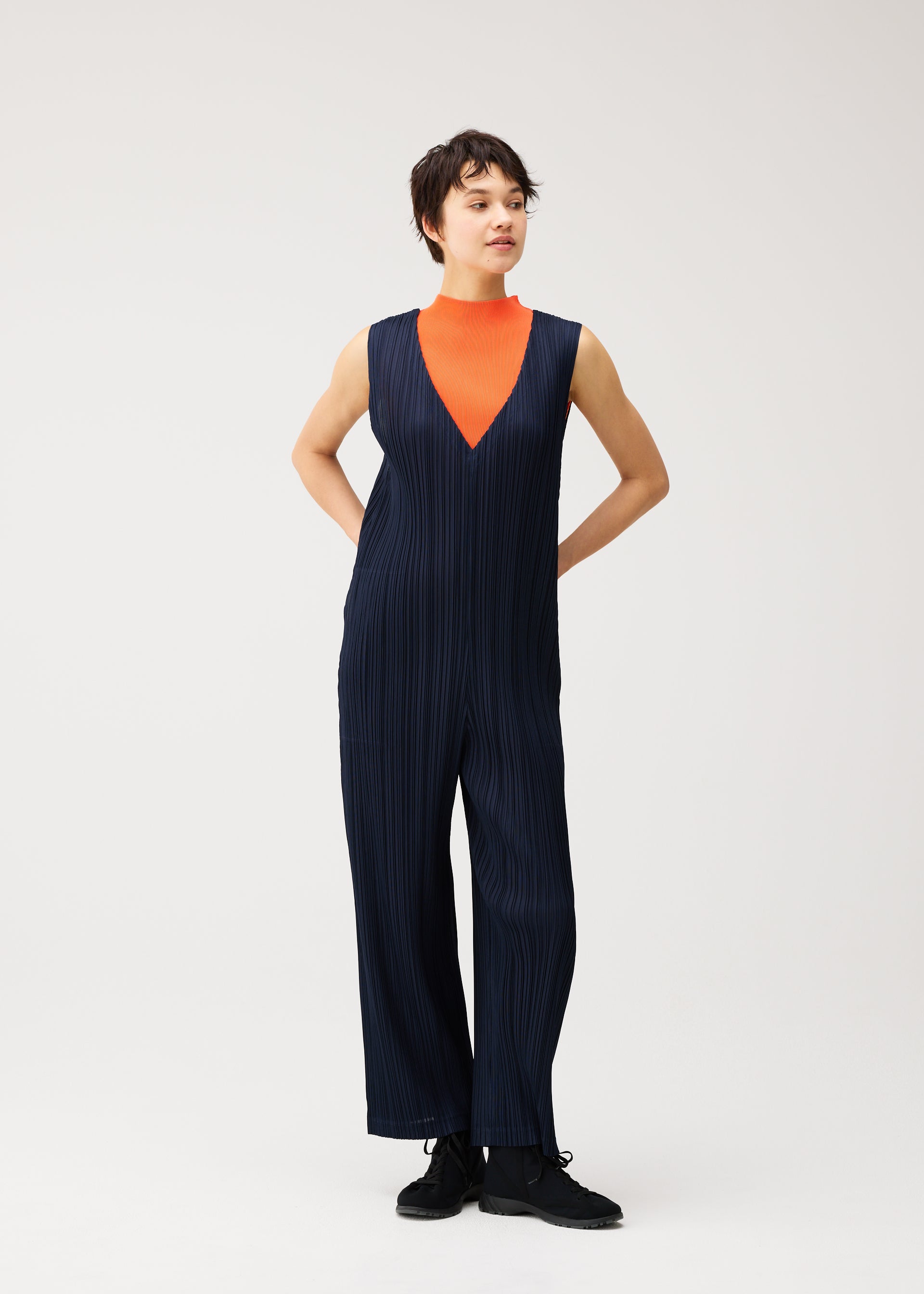 Zoomed view A person stands with hands behind their back against a plain light background, wearing the PLEATS PLEASE ISSEY MIYAKE THICKER BOTTOMS 2—a sleeveless navy blue V-neck jumpsuit featuring pockets and a sharply pleated front, layered over an orange turtleneck. Crafted from thick fabric, this structured outfit exudes modern sophistication.