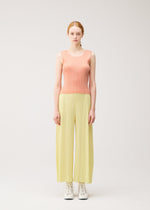 A person with fair skin and red hair faces forward, wearing the MONTHLY COLORS : JULY wide-legged yellow pleated pants by PLEATS PLEASE ISSEY MIYAKE, a peach round-neck tank top, and white lace-up shoes against a plain light background.