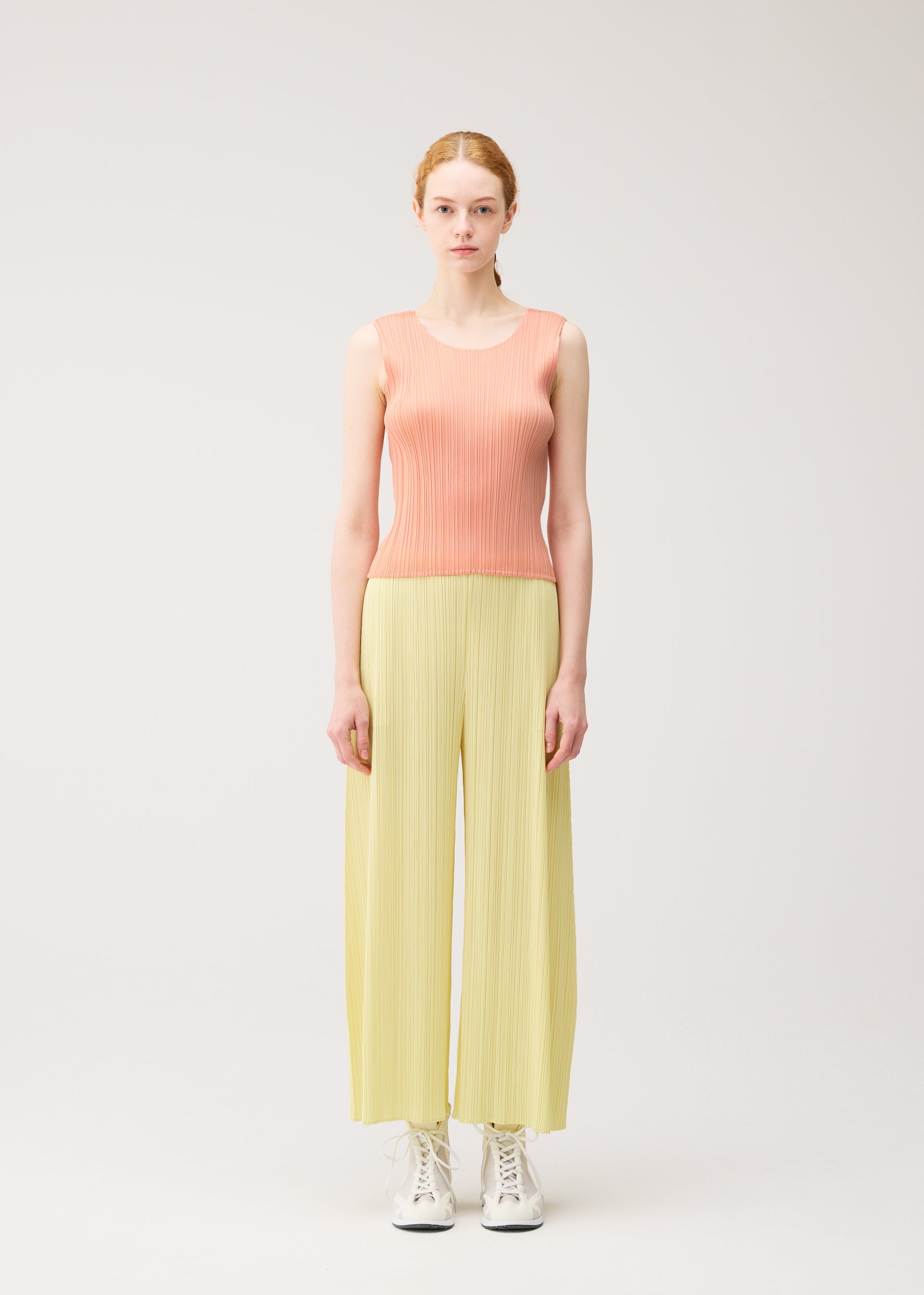 Zoomed view A person with fair skin and red hair faces forward, wearing the MONTHLY COLORS : JULY wide-legged yellow pleated pants by PLEATS PLEASE ISSEY MIYAKE, a peach round-neck tank top, and white lace-up shoes against a plain light background.