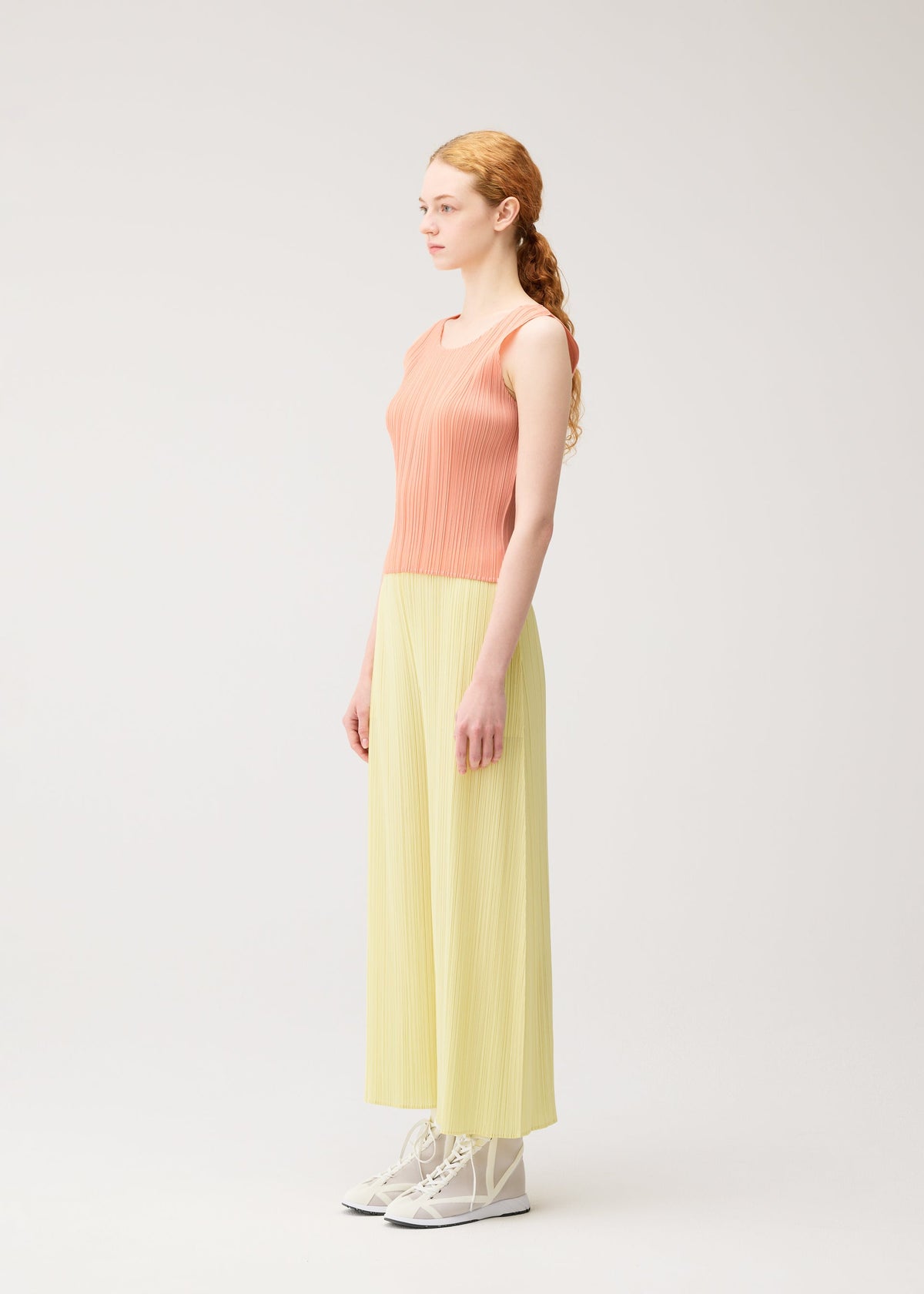 A woman with long red hair in a ponytail stands sideways, wearing PLEATS PLEASE ISSEY MIYAKE MONTHLY COLORS: JULY—coral sleeveless top and light yellow pleated pants—with white ankle boots against a plain light background. view 8 of 10