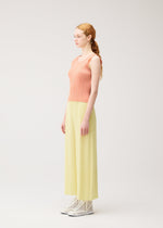 A woman with long red hair in a ponytail stands sideways, wearing PLEATS PLEASE ISSEY MIYAKE MONTHLY COLORS: JULY—coral sleeveless top and light yellow pleated pants—with white ankle boots against a plain light background.