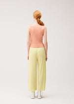 A person with reddish hair in a low braid wears the MONTHLY COLORS : JULY sleeveless pleated shirt and pale yellow pleated pants by PLEATS PLEASE ISSEY MIYAKE, standing with their back to the camera against a plain light background.