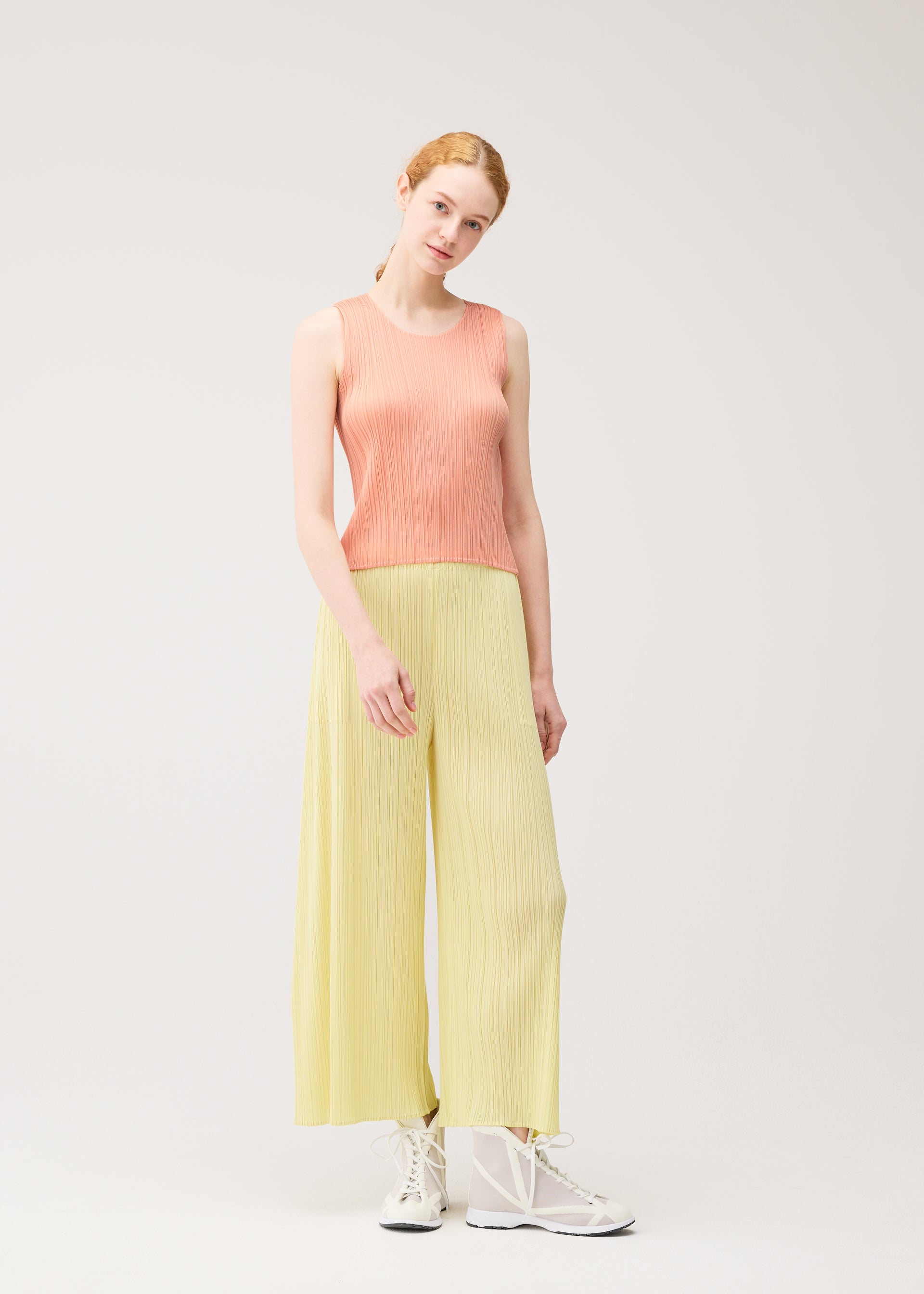 Zoomed view A woman with light skin and red hair models the MONTHLY COLORS : JULY sleeveless shirt and wide-leg pants by PLEATS PLEASE ISSEY MIYAKE, paired with white high-top sneakers against a plain light background.