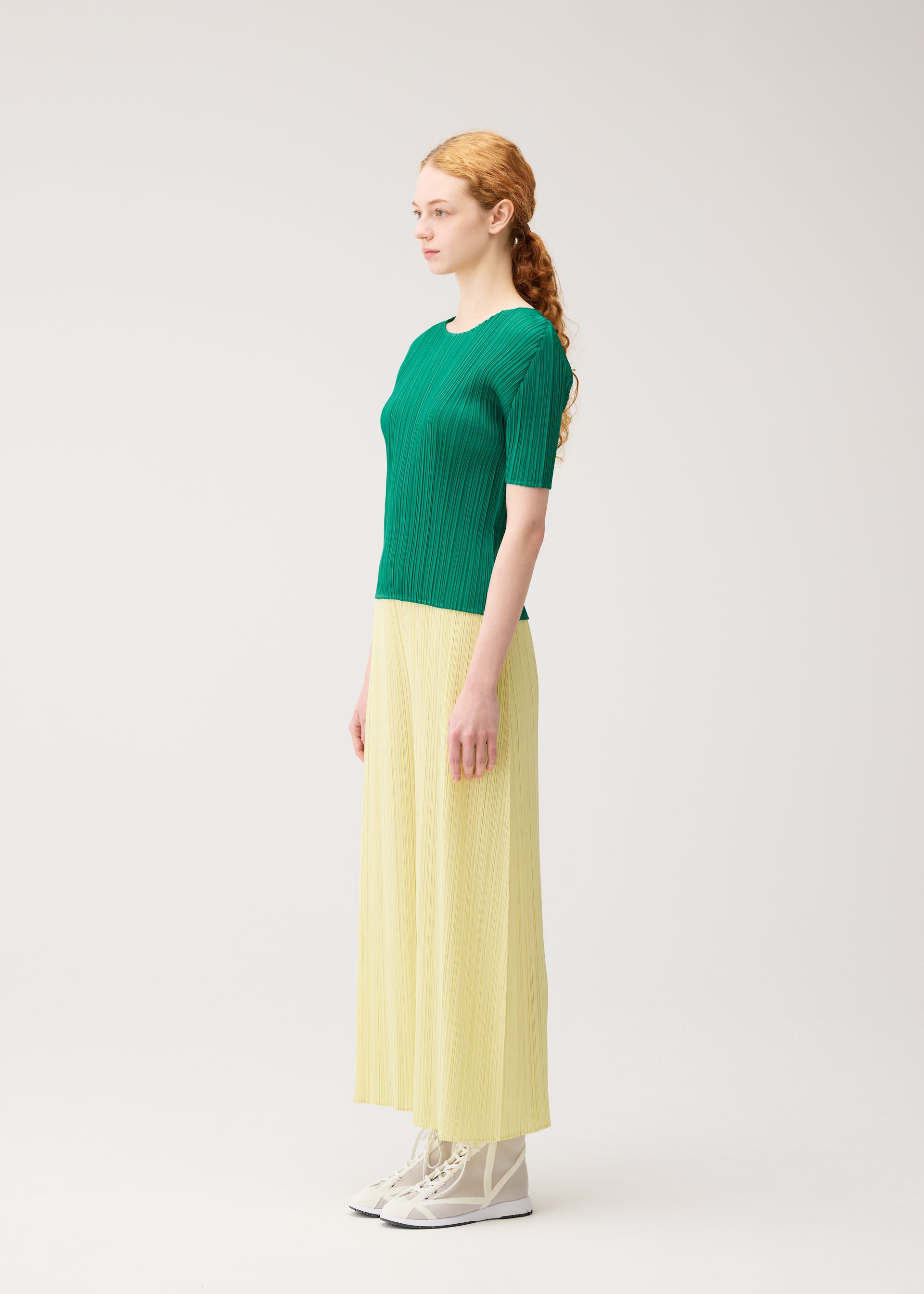 Zoomed view A woman with long red hair in a ponytail stands in profile against a plain background, wearing the MONTHLY COLORS : JULY collection by PLEATS PLEASE ISSEY MIYAKE—a green top, flowing yellow skirt, and vibrant white lace-up shoes.