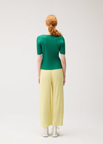 A person with long red hair in a low ponytail stands facing away, wearing PLEATS PLEASE ISSEY MIYAKE MONTHLY COLORS : JULY—a green top and wide-leg, pale yellow pleated pants—paired with white shoes against a plain light background.