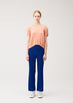 A person with light skin and reddish-blonde hair tied back wears the MONTHLY COLORS : JULY blue pleated pants by PLEATS PLEASE ISSEY MIYAKE, paired with a peach round-neck top and white sneakers, standing against a plain background.