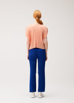 A person with long, braided reddish hair faces away, wearing the MONTHLY COLORS : JULY loose pleated top and blue pants by PLEATS PLEASE ISSEY MIYAKE, paired with white shoes against a light background.