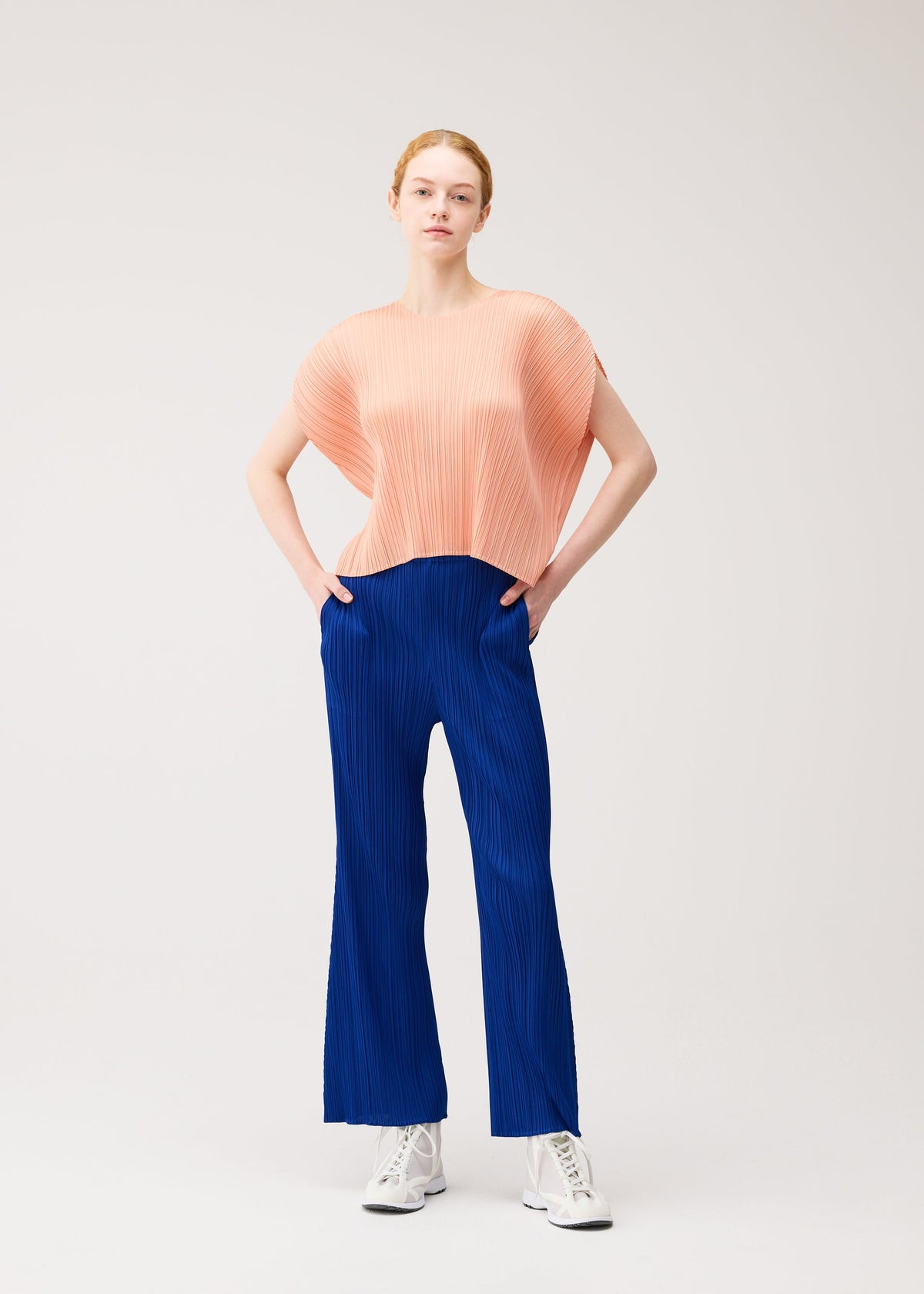 A woman wears PLEATS PLEASE ISSEY MIYAKE’s MONTHLY COLORS : JULY—a loose peach top with bright blue pleated pants and white sneakers—posing confidently against a light background with her hands in her pockets. view 11 of 11