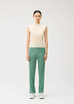 A woman stands facing forward against a plain light background, wearing the MONTHLY COLORS : AUGUST sleeveless cream high-neck top and green pleated pants with straight fold lines from PLEATS PLEASE ISSEY MIYAKE, paired with white sneakers.