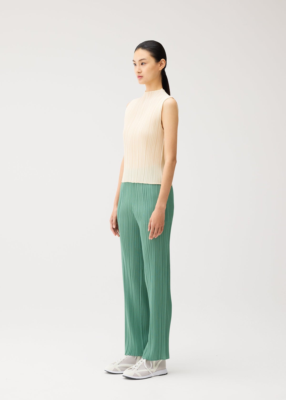 Wearing the “MONTHLY COLORS : AUGUST” collection from PLEATS PLEASE ISSEY MIYAKE, a woman stands in profile against a plain light background. She wears a sleeveless beige high-neck top paired with loose green pleated pants featuring straight fold lines, and completes the look with white sneakers. view 8 of 10