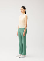 Wearing the “MONTHLY COLORS : AUGUST” collection from PLEATS PLEASE ISSEY MIYAKE, a woman stands in profile against a plain light background. She wears a sleeveless beige high-neck top paired with loose green pleated pants featuring straight fold lines, and completes the look with white sneakers.