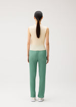 A person with long dark hair in a ponytail stands facing away, wearing the MONTHLY COLORS : AUGUST sleeveless cream high-neck top and green pleated pants with straight fold lines from PLEATS PLEASE ISSEY MIYAKE, paired with white shoes, against a plain light background.