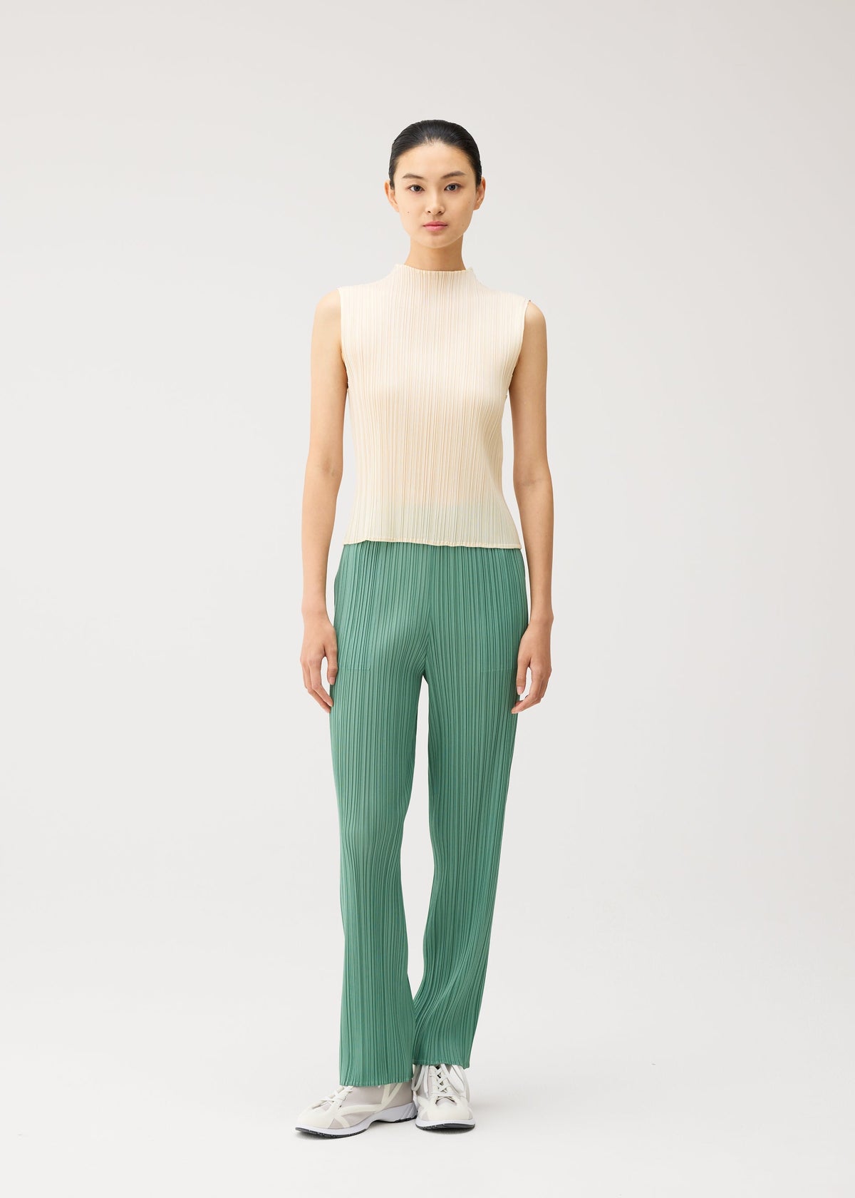 A woman stands against a plain background, wearing the MONTHLY COLORS : AUGUST sleeveless high-neck top and green pleated pants with straight fold lines from PLEATS PLEASE ISSEY MIYAKE, paired with white sneakers. Her straight black hair is pulled back, and she has a neutral expression. view 10 of 10