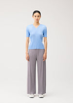 A woman stands facing forward, wearing the MONTHLY COLORS : AUGUST light blue short-sleeved V-neck top with pleated detailing and wide-legged gray pleated pants by PLEATS PLEASE ISSEY MIYAKE, paired with white sneakers against a plain light background.