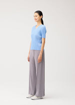 A woman stands in a studio wearing the MONTHLY COLORS : AUGUST collection from PLEATS PLEASE ISSEY MIYAKE—a light blue, short-sleeved V-neck top paired with wide-leg gray pleated pants and white shoes. She looks ahead, smiling slightly against a plain, light background—an ensemble that captures the essence of midsummer style.