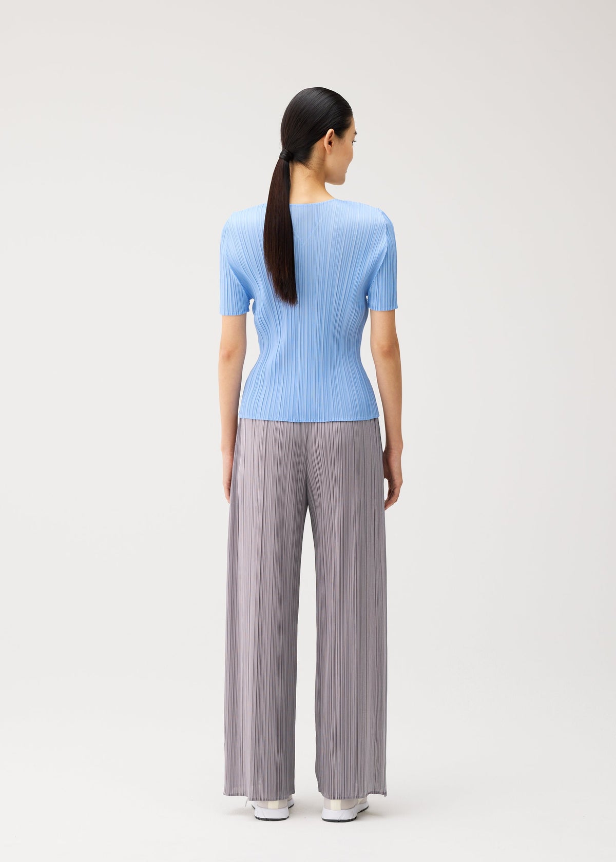 A woman with long dark hair in a ponytail stands facing away, wearing pieces from the MONTHLY COLORS : AUGUST collection by PLEATS PLEASE ISSEY MIYAKE—a light blue, short-sleeved V-neck top paired with loose gray pleated pants and white shoes—on a plain light background. This ensemble is perfect for a relaxed midsummer day. view 9 of 10