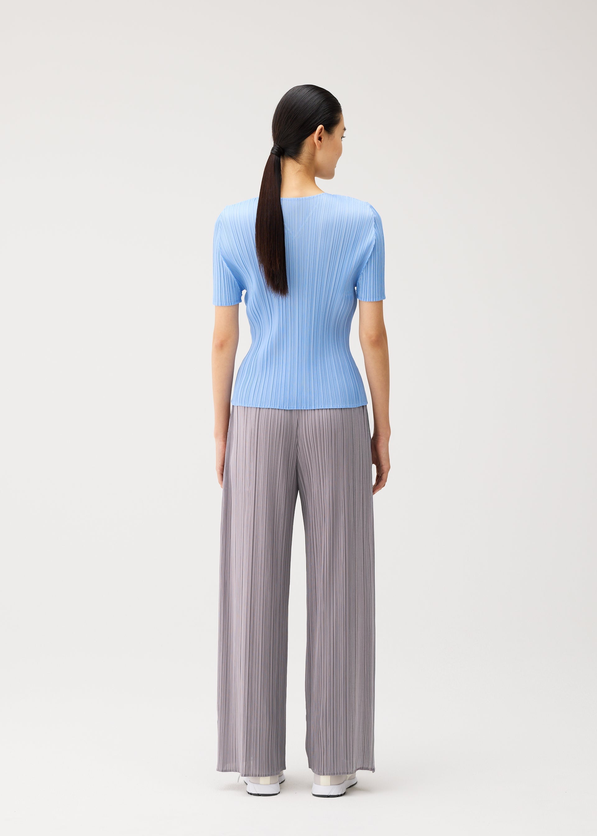 Zoomed view A woman with long dark hair in a ponytail stands facing away, wearing pieces from the MONTHLY COLORS : AUGUST collection by PLEATS PLEASE ISSEY MIYAKE—a light blue, short-sleeved V-neck top paired with loose gray pleated pants and white shoes—on a plain light background. This ensemble is perfect for a relaxed midsummer day.