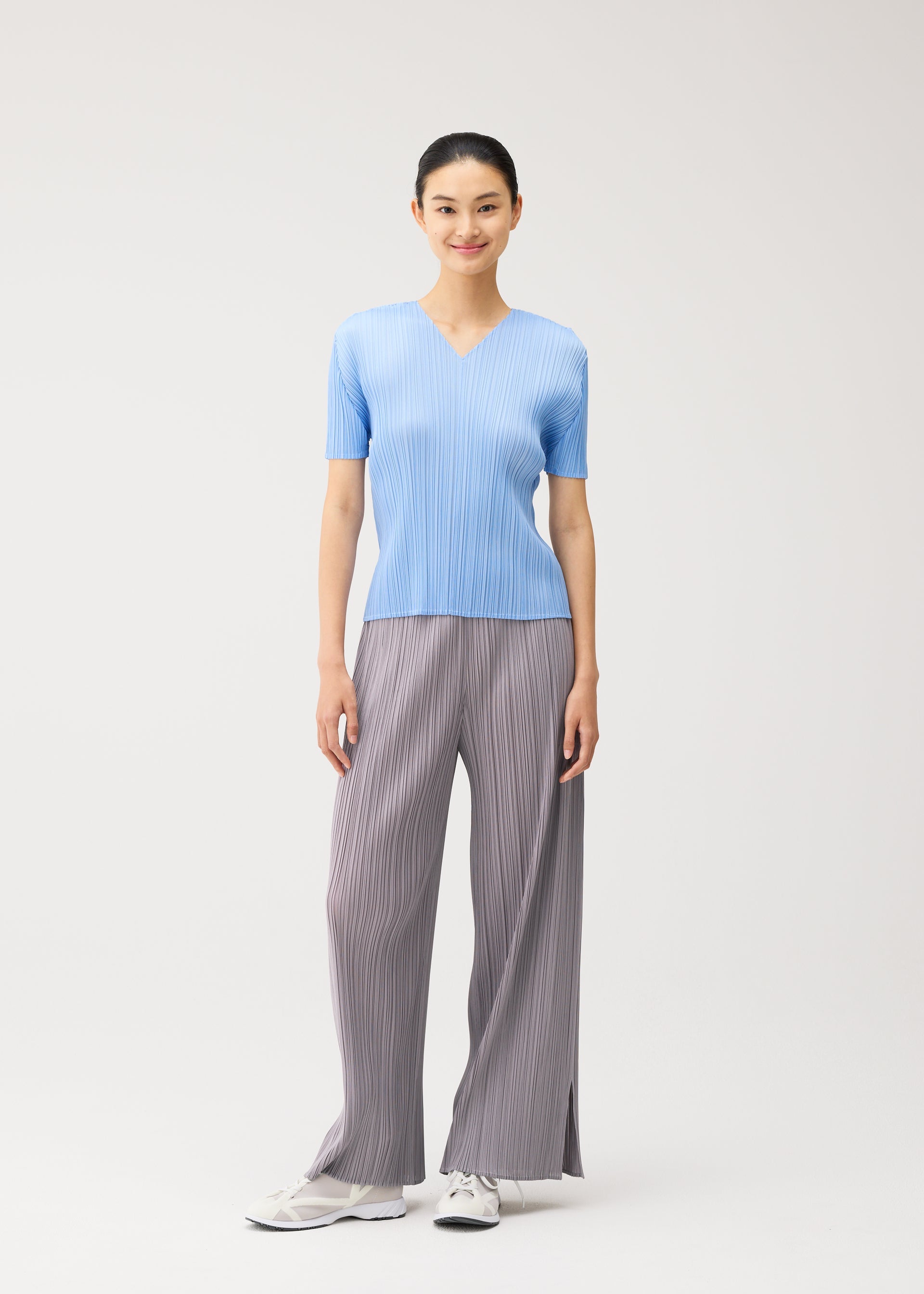 Zoomed view A woman stands against a plain background, smiling at the camera in a light blue, short-sleeved V-neck top and wide gray pleated pants from the MONTHLY COLORS : AUGUST collection by PLEATS PLEASE ISSEY MIYAKE, paired with white sneakers—her look ideal for a midsummer day.