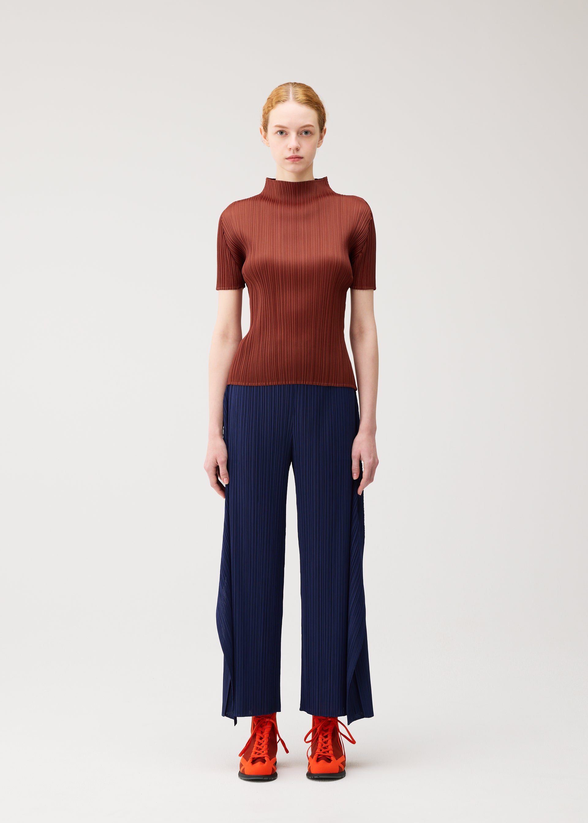 Zoomed view A woman stands against a plain background, wearing the fitted, short-sleeved “MONTHLY COLORS : SEPTEMBER” top by PLEATS PLEASE ISSEY MIYAKE in a warm brown shade that highlights seasonal colors. She pairs it with navy wide-leg pants featuring slits and bright orange lace-up shoes.