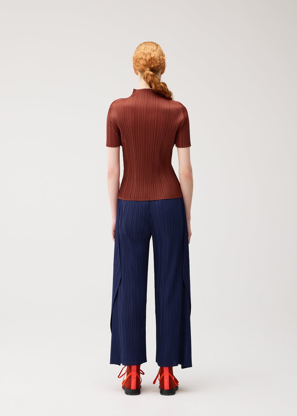 A person with long red hair in a braid faces away, dressed in the MONTHLY COLORS : SEPTEMBER collection from PLEATS PLEASE ISSEY MIYAKE—a fitted ribbed brown high-neck top styled with navy wide-leg pleated pants and red heeled shoes with black soles—standing against a plain light background. The ensemble showcases rich seasonal colors that enhance the overall look. view 9 of 10