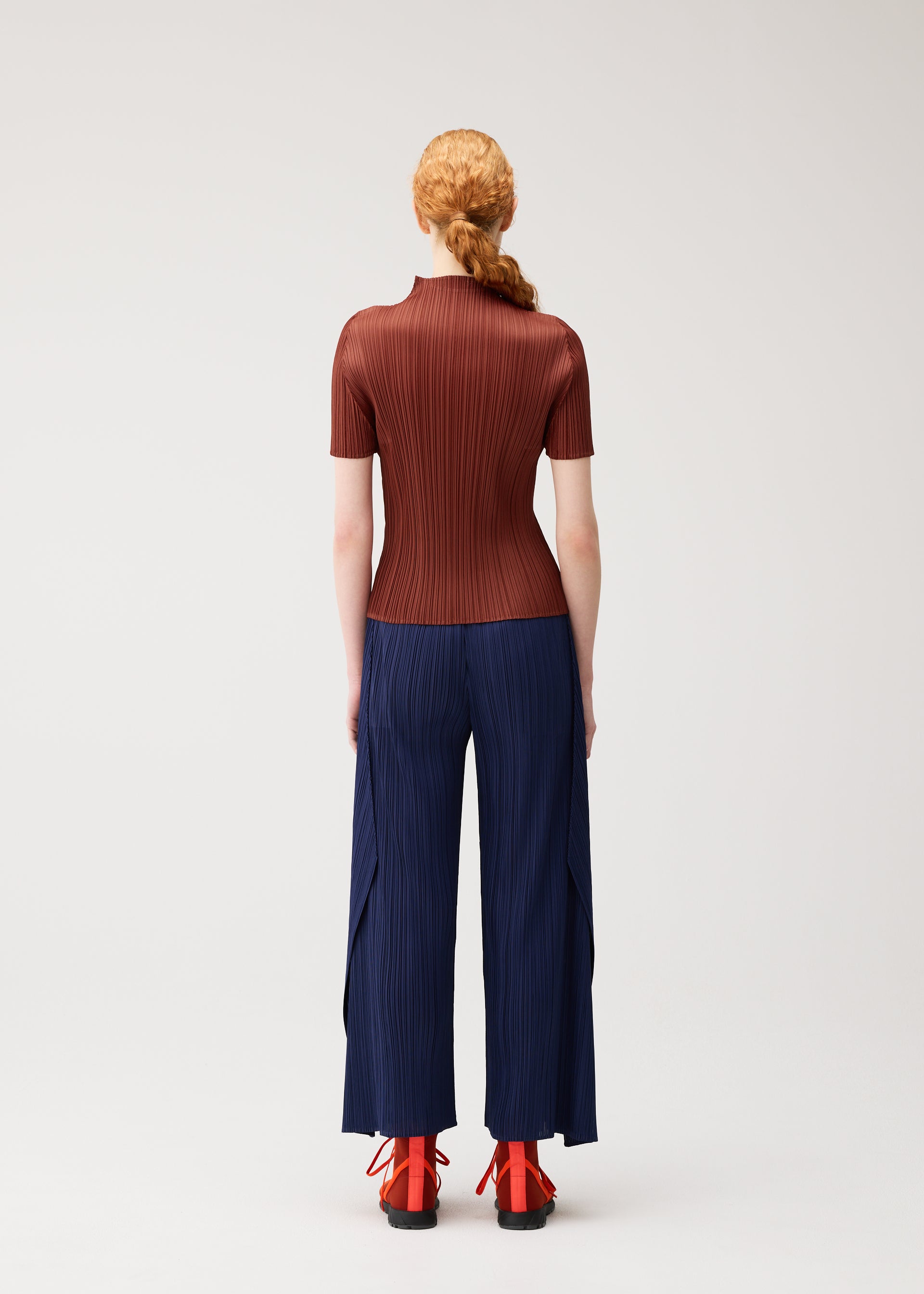 Zoomed view A person with long red hair in a braid faces away, dressed in the MONTHLY COLORS : SEPTEMBER collection from PLEATS PLEASE ISSEY MIYAKE—a fitted ribbed brown high-neck top styled with navy wide-leg pleated pants and red heeled shoes with black soles—standing against a plain light background. The ensemble showcases rich seasonal colors that enhance the overall look.