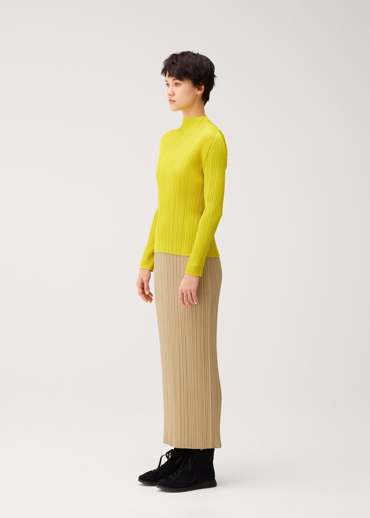 Person standing in profile against a plain background, wearing the MONTHLY COLORS : OCTOBER by PLEATS PLEASE ISSEY MIYAKE—a bright yellow long-sleeved, high-neck top and beige pleated skirt with black shoes. view 8 of 10